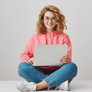 copywriter ready to work. attractive smart curly haired woman in trendy glasses sitting on floor, holding laptop on laps and smiling at camera, writing post in social network or editing text